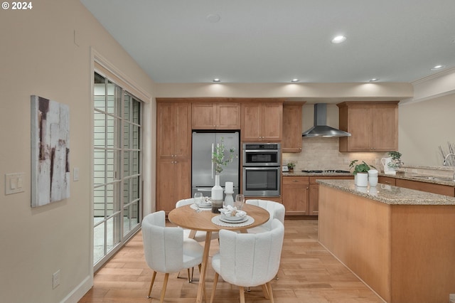 kitchen featuring light stone countertops, stainless steel appliances, wall chimney range hood, and light hardwood / wood-style flooring