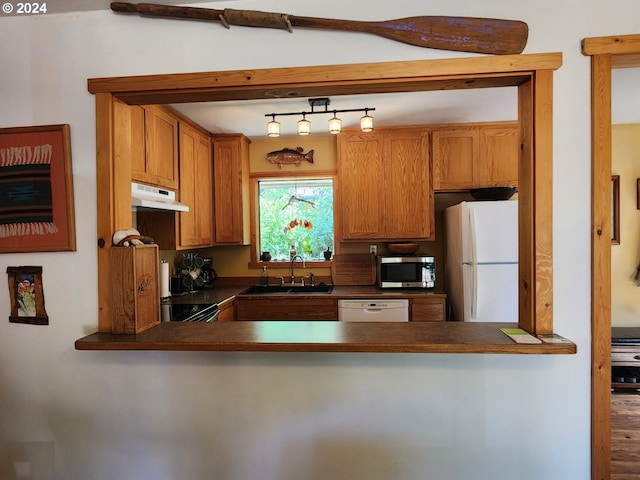 kitchen featuring under cabinet range hood, a peninsula, white appliances, a sink, and dark countertops