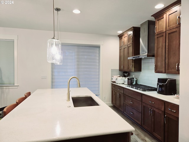 kitchen featuring wall chimney exhaust hood, a kitchen island with sink, stainless steel gas cooktop, sink, and pendant lighting