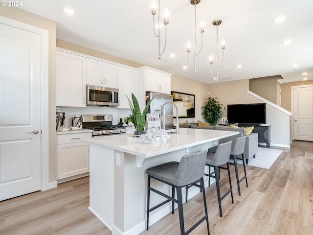 kitchen featuring appliances with stainless steel finishes, light wood-type flooring, a kitchen island with sink, decorative light fixtures, and white cabinets