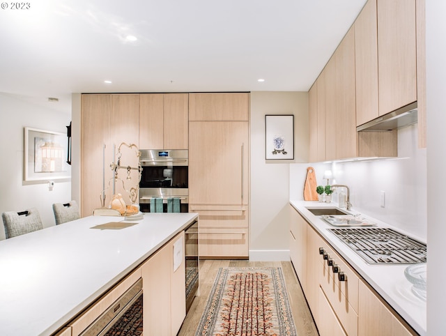 kitchen featuring stainless steel double oven, under cabinet range hood, a sink, light countertops, and light brown cabinetry