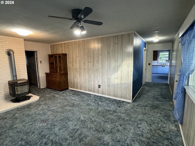 unfurnished living room with a wood stove, ceiling fan, dark carpet, and ornamental molding