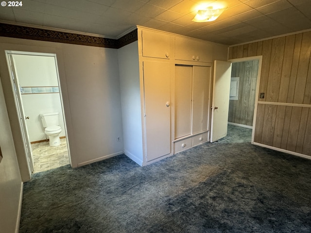 unfurnished bedroom with dark colored carpet, a closet, ornamental molding, wooden walls, and ensuite bath