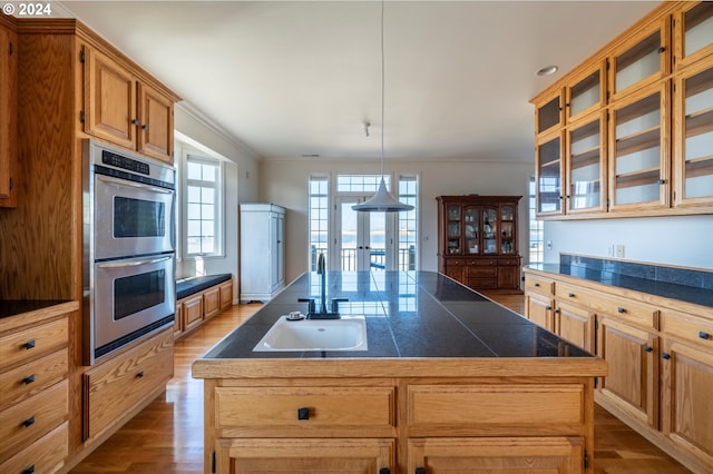 kitchen with an island with sink, ornamental molding, wood finished floors, a sink, and stainless steel double oven