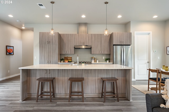 kitchen with dark wood-type flooring, hanging light fixtures, a center island with sink, stainless steel refrigerator, and a kitchen breakfast bar