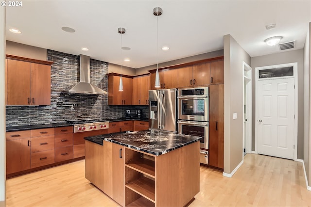 kitchen with appliances with stainless steel finishes, wall chimney range hood, light hardwood / wood-style floors, a kitchen island, and decorative backsplash