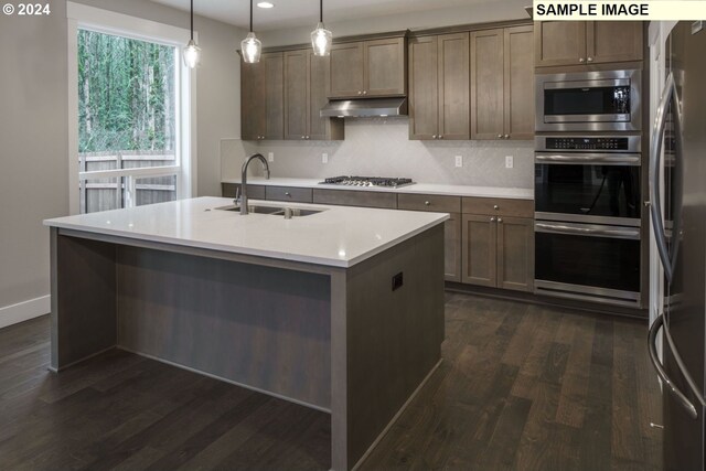 kitchen with dark wood-type flooring, a kitchen island with sink, appliances with stainless steel finishes, sink, and pendant lighting