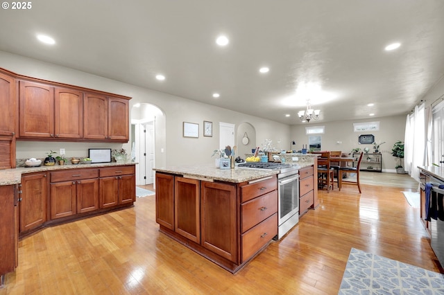 kitchen with stainless steel range oven, light stone counters, hanging light fixtures, a center island with sink, and light hardwood / wood-style flooring