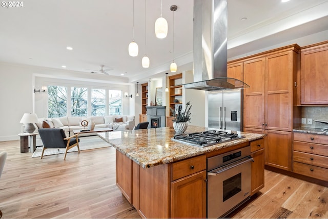 kitchen with appliances with stainless steel finishes, brown cabinets, open floor plan, decorative light fixtures, and island exhaust hood