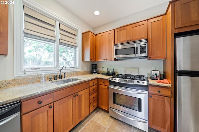 kitchen with a sink, appliances with stainless steel finishes, stone finish floor, light stone countertops, and brown cabinetry