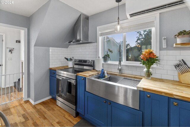 kitchen featuring wall chimney range hood, stainless steel electric stove, pendant lighting, wooden counters, and a wall mounted AC