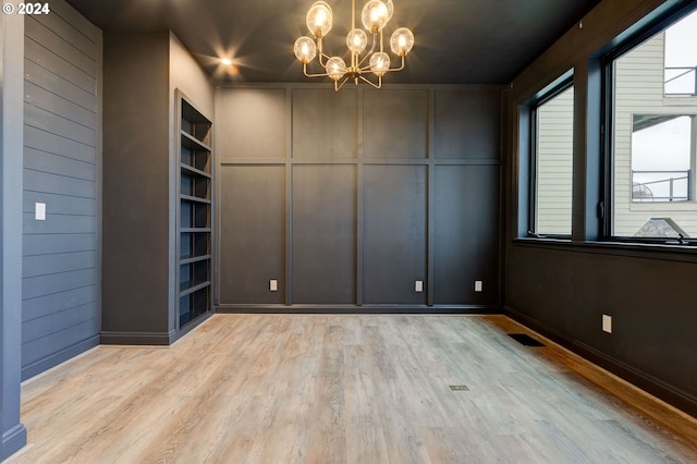 empty room with light hardwood / wood-style flooring, wooden walls, built in features, and an inviting chandelier