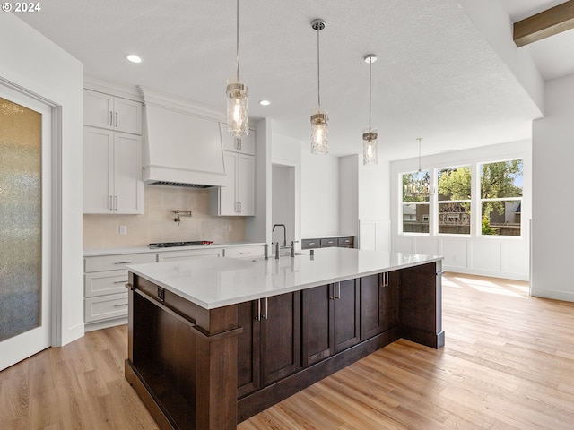 kitchen with decorative light fixtures, light hardwood / wood-style flooring, a kitchen island with sink, and custom exhaust hood