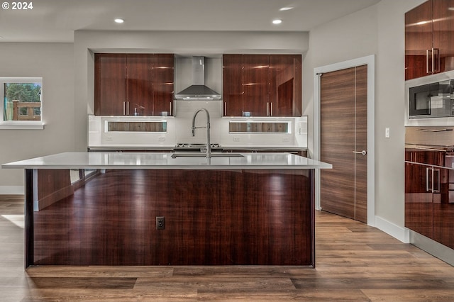 kitchen featuring a kitchen island with sink, wall chimney range hood, stainless steel microwave, backsplash, and light hardwood / wood-style floors