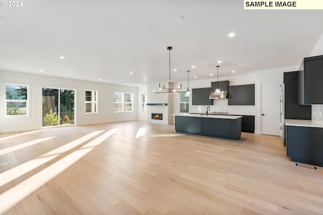 kitchen with decorative light fixtures, backsplash, an island with sink, wall chimney range hood, and light wood-type flooring