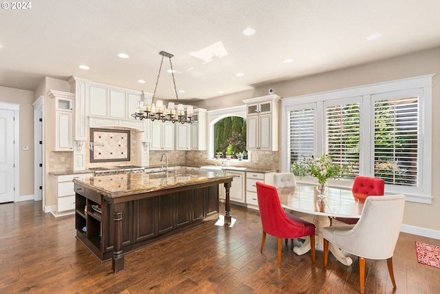 kitchen with backsplash, light stone countertops, an island with sink, decorative light fixtures, and dark hardwood / wood-style flooring