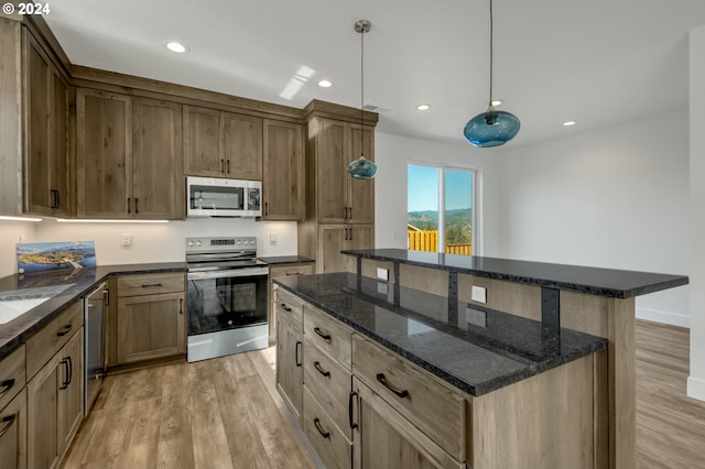 kitchen with appliances with stainless steel finishes, light wood-type flooring, a center island, dark stone counters, and pendant lighting