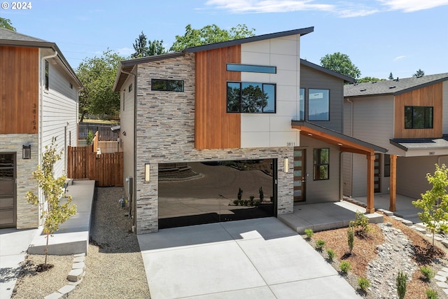 contemporary home with fence, concrete driveway, a patio area, stone siding, and an attached garage