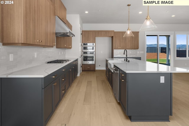 kitchen with light stone counters, an island with sink, pendant lighting, stainless steel appliances, and light hardwood / wood-style floors