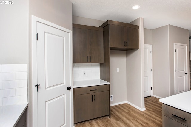 kitchen featuring light countertops, light wood-style flooring, decorative backsplash, dark brown cabinets, and baseboards