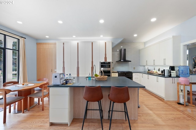 kitchen featuring white cabinets, wall chimney range hood, light hardwood / wood-style flooring, decorative backsplash, and gas stove