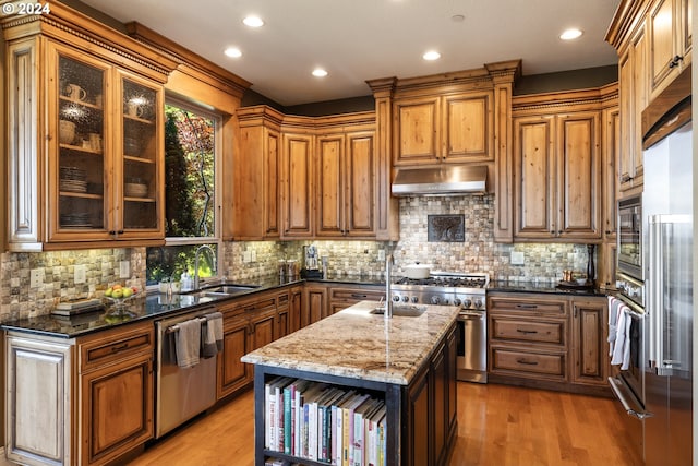 kitchen featuring light hardwood / wood-style flooring, built in appliances, decorative backsplash, dark stone countertops, and an island with sink