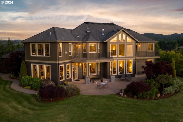 back house at dusk with a patio, a balcony, and a lawn