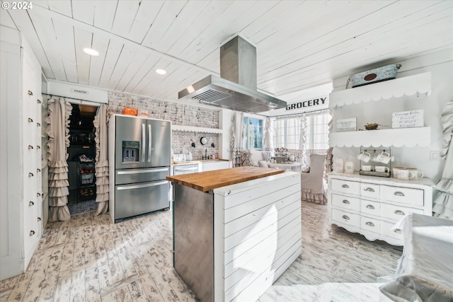 kitchen featuring white cabinets, stainless steel refrigerator with ice dispenser, island range hood, butcher block counters, and brick wall