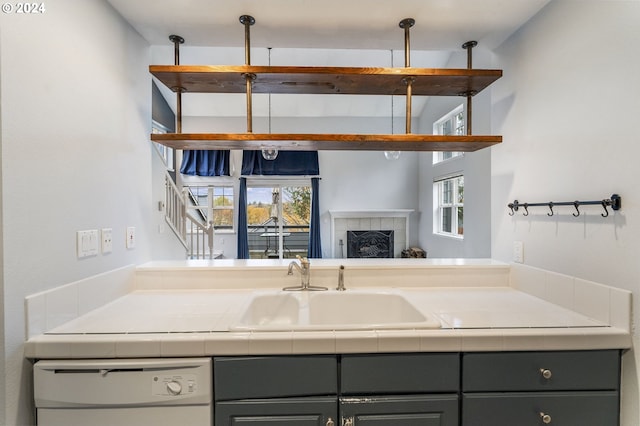 kitchen with dishwasher, tile countertops, gray cabinetry, a tile fireplace, and sink