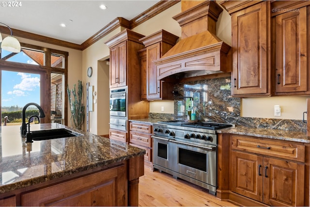 kitchen featuring stainless steel appliances, custom range hood, crown molding, light wood-type flooring, and dark stone countertops