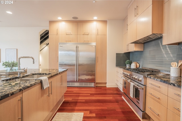 kitchen with high end appliances, dark wood-type flooring, sink, light brown cabinets, and dark stone countertops