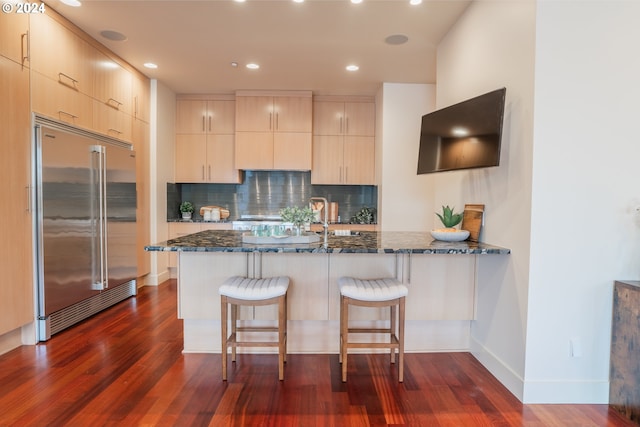 kitchen featuring dark wood-type flooring, built in refrigerator, dark stone countertops, tasteful backsplash, and kitchen peninsula