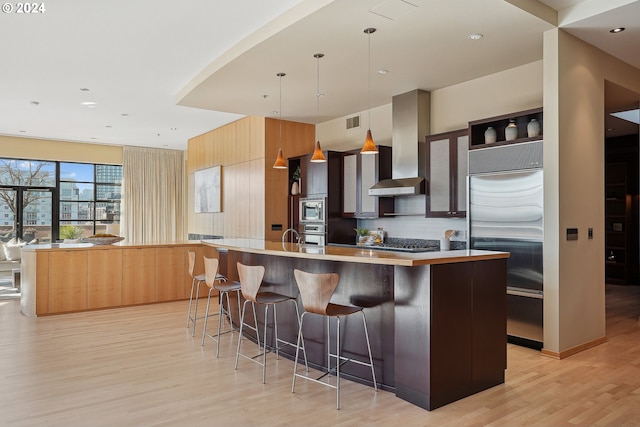 kitchen with a kitchen bar, wall chimney exhaust hood, built in appliances, decorative light fixtures, and a center island with sink