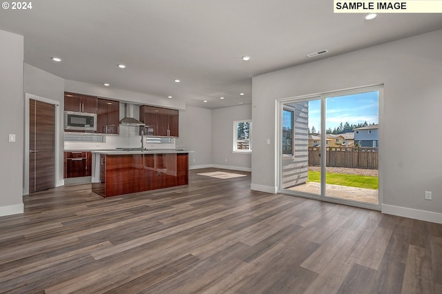 kitchen featuring a kitchen island with sink, stainless steel microwave, wall chimney exhaust hood, dark wood-type flooring, and a breakfast bar