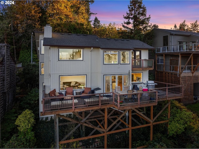 back of house at dusk featuring a shingled roof, a balcony, and a chimney