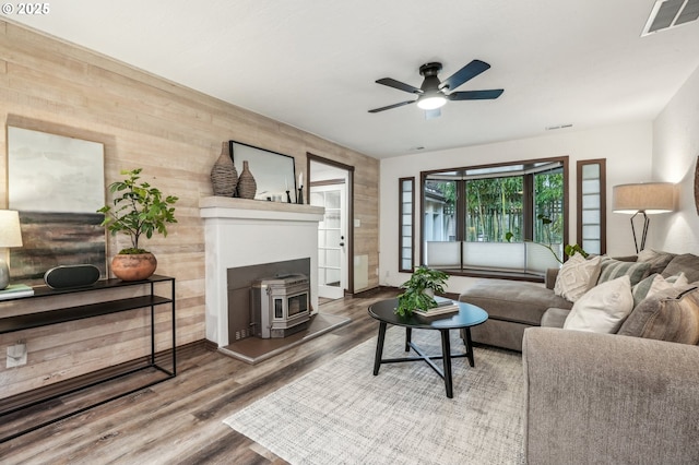 living room with ceiling fan, wood-type flooring, wooden walls, and a wood stove