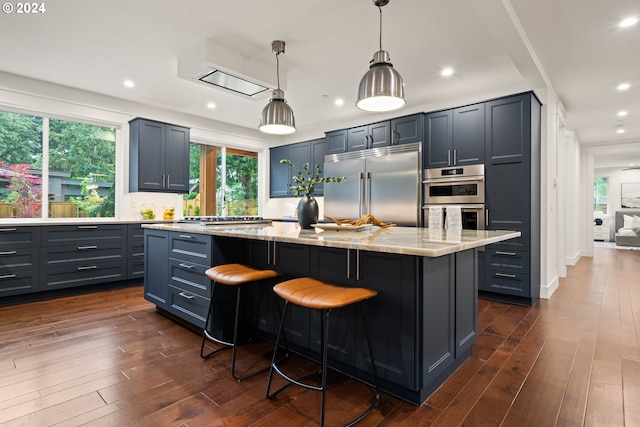 kitchen with light stone countertops, appliances with stainless steel finishes, decorative light fixtures, a kitchen island, and dark hardwood / wood-style flooring