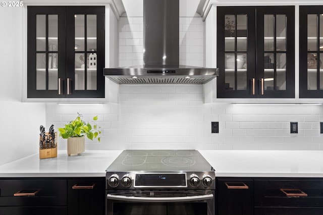 kitchen with stainless steel electric range oven, glass insert cabinets, dark cabinetry, and wall chimney exhaust hood