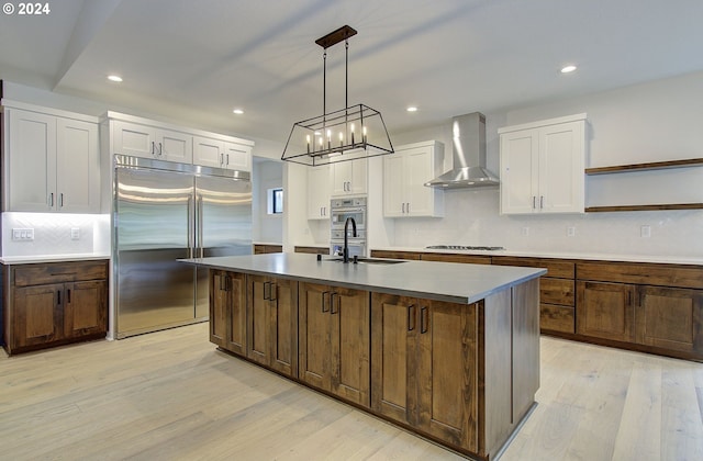 kitchen with appliances with stainless steel finishes, wall chimney exhaust hood, decorative light fixtures, light hardwood / wood-style flooring, and an island with sink