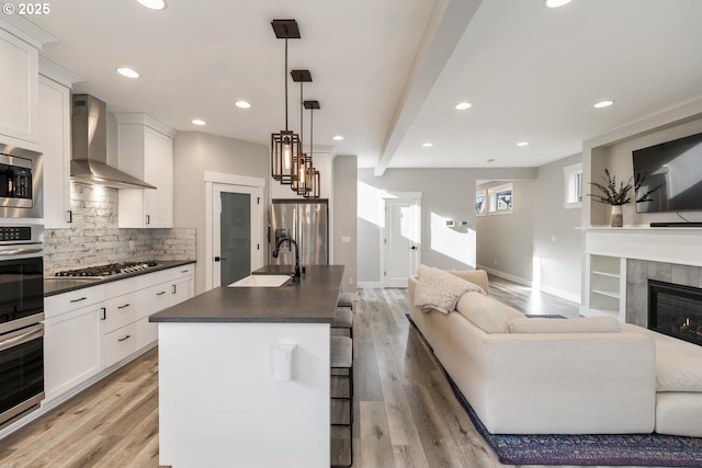 kitchen with appliances with stainless steel finishes, decorative light fixtures, an island with sink, sink, and wall chimney range hood