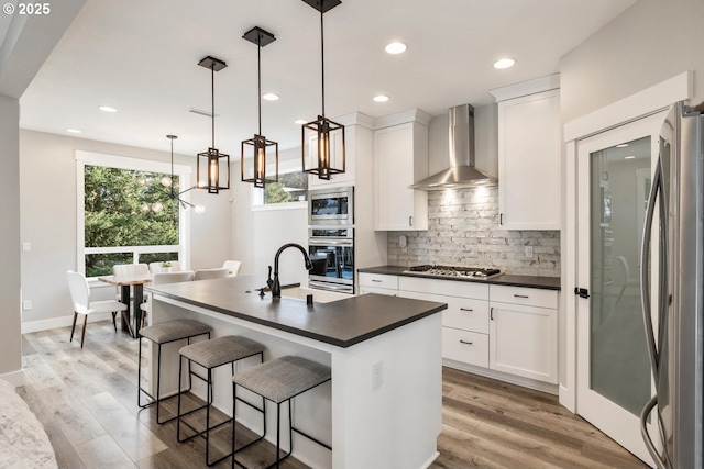 kitchen featuring an island with sink, white cabinetry, hanging light fixtures, stainless steel appliances, and wall chimney range hood