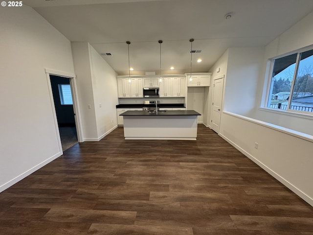 kitchen with dark countertops, stainless steel microwave, hanging light fixtures, white cabinets, and a kitchen island with sink