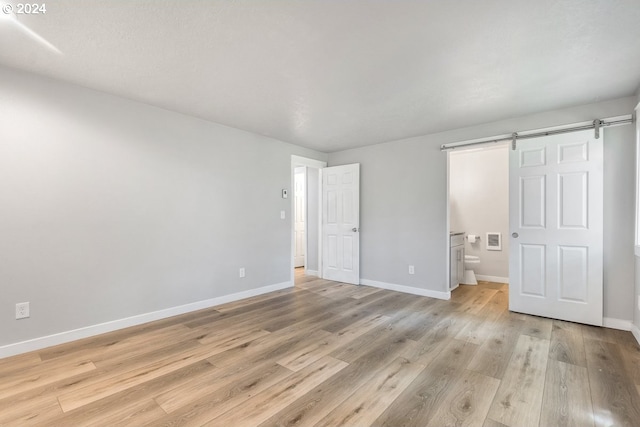 unfurnished bedroom featuring a barn door, baseboards, light wood-type flooring, and ensuite bath