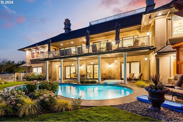 back house at dusk featuring a balcony, a patio area, and a pool with hot tub