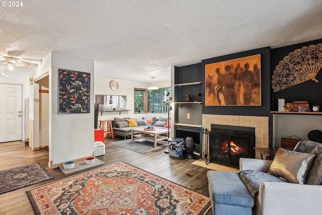 living room with ceiling fan, a tiled fireplace, a textured ceiling, and hardwood / wood-style floors