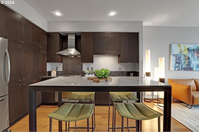 kitchen with light stone countertops, a breakfast bar area, light hardwood / wood-style floors, wall chimney exhaust hood, and stainless steel refrigerator