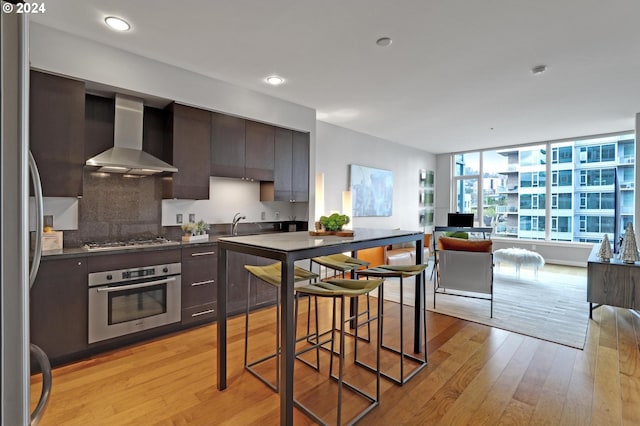 kitchen with dark brown cabinets, backsplash, light hardwood / wood-style flooring, stainless steel appliances, and wall chimney exhaust hood