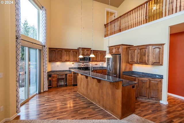 kitchen featuring a towering ceiling, stainless steel appliances, dark wood-type flooring, hanging light fixtures, and an island with sink