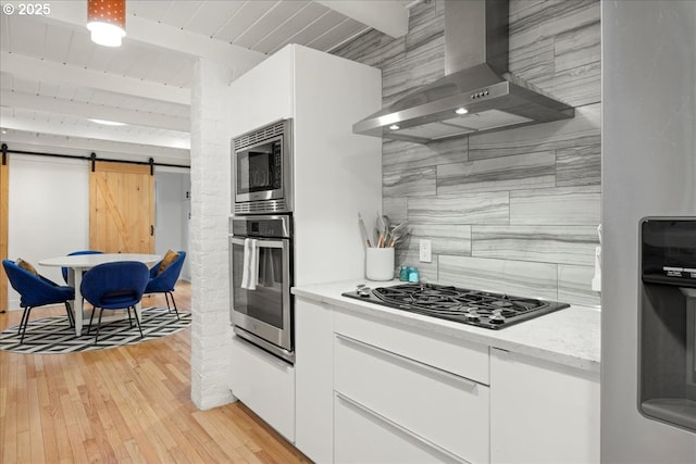 kitchen featuring wall chimney range hood, appliances with stainless steel finishes, white cabinetry, a barn door, and beamed ceiling