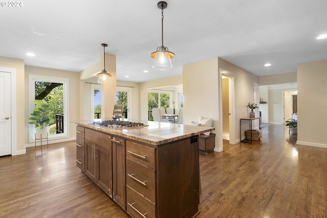 kitchen with stainless steel gas stovetop, hanging light fixtures, dark hardwood / wood-style floors, a kitchen island, and light stone counters